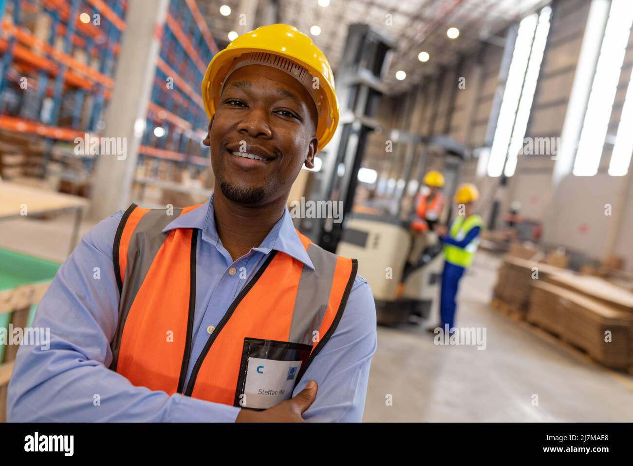 Ritratto di sorridente giovane uomo sicuro indossando un elmetto con le braccia incrociate in piedi in magazzino Foto Stock