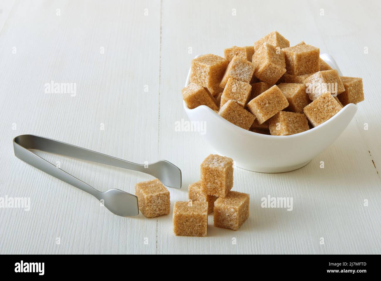 Zucchero di canna a forma di cubo in ciotola di porcellana su un tavolo di legno bianco con pinze di zucchero d'argento. Primo piano, nessuna persona. Foto Stock