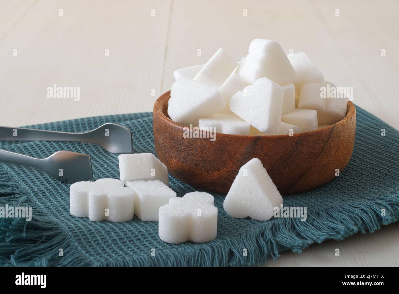 Cubetti di zucchero a forma di ponte in ciotola di legno su un tovagliolo turchese con pinze di zucchero d'argento. Primo piano, tavolo bianco in legno, niente persone. Foto Stock