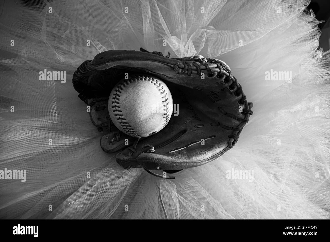 Guanto da baseball con softball a metà, seduto in cima a un tutu balletto bianco. Foto Stock