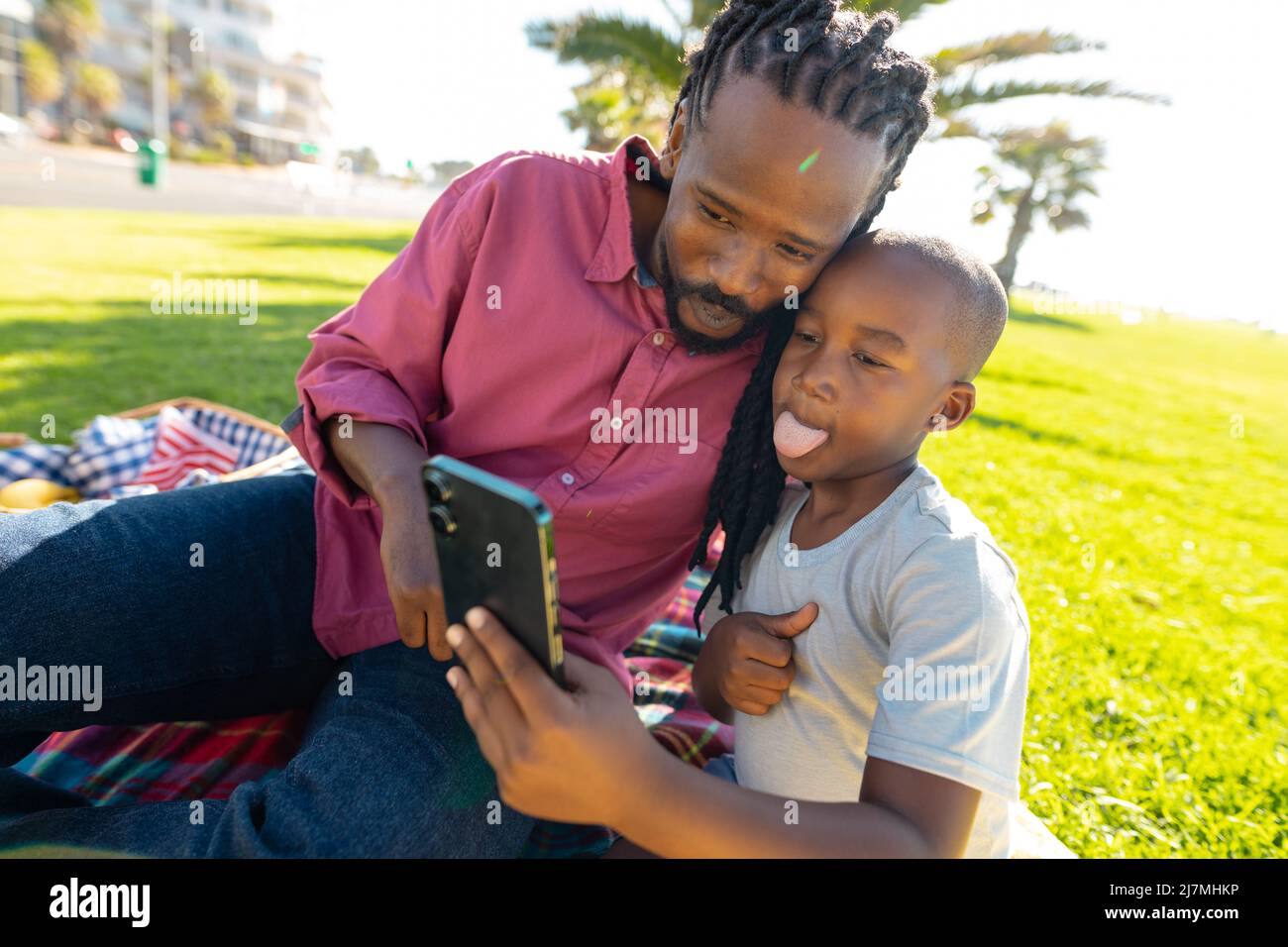 Ragazzo afroamericano che attacca la lingua mentre prende selfie con il padre al parco in giorno di sole Foto Stock
