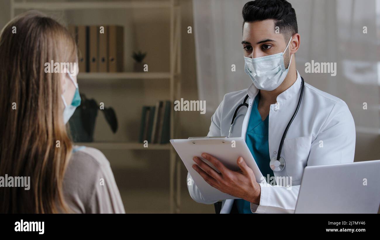 Medico maschio arabo in maschera medica fare note sulla clipboard consulenza paziente femmina spiegare risultati medici dando prescrizione Foto Stock