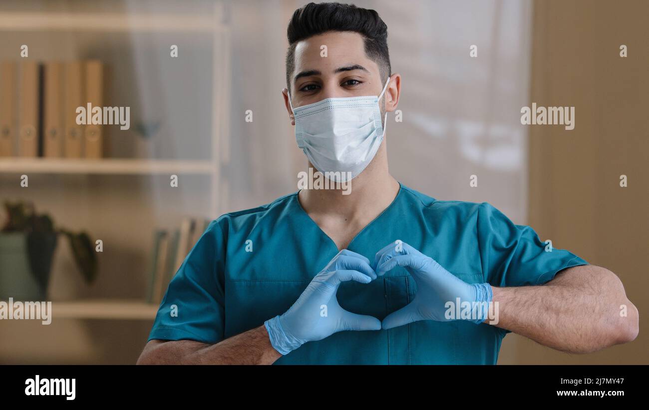 Ritratto uomo giovane medico arabo infermiere indossare maschera viso uniforme medica mostrando a fotocamera cuore amore segno gesto uomo mani in guanti simbolizzazione Foto Stock