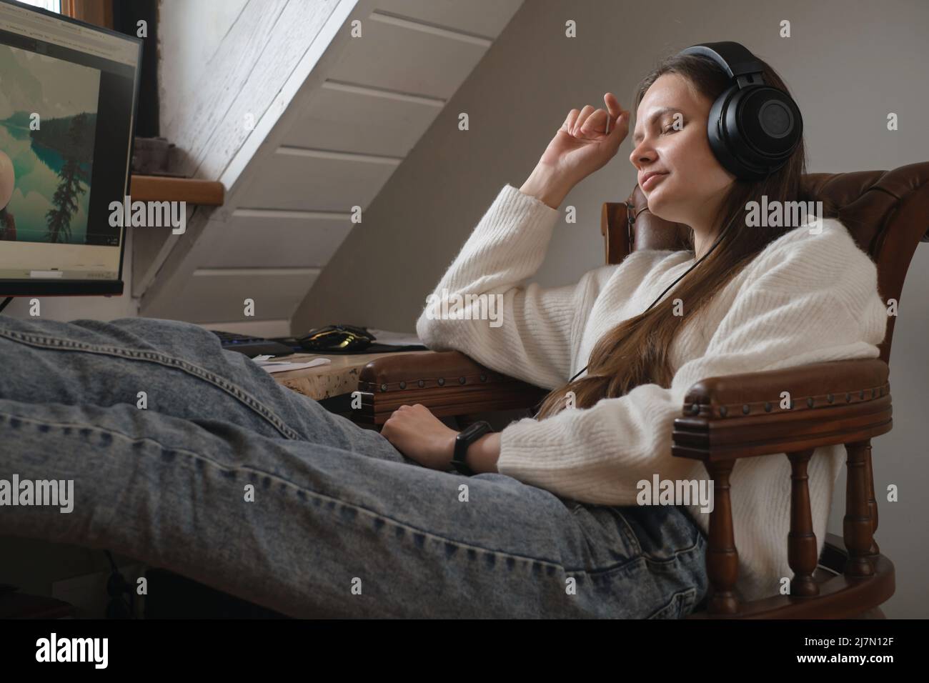 Giovane donna che ascolta la musica in grandi cuffie vicino al computer a casa. Creazione di una lista di gioco, godendo di tempo libero o di lavoro. Femmina al suo 20s vicino Foto Stock