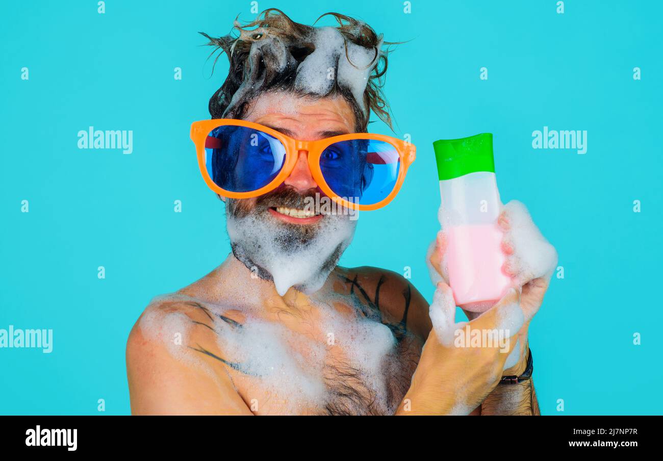 Uomo divertente sotto la doccia con una bottiglia di shampoo. Ragazzo bearded in grandi vetri con schiuma sulla testa. Lavaggio dei capelli. Foto Stock