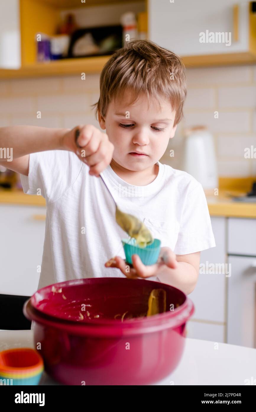 Il capretto piccolo 5 anni cuoca i cupcakes nella cucina leggera in casa. Il ragazzo piccolo usa gli stampi di cottura del silicone Foto Stock