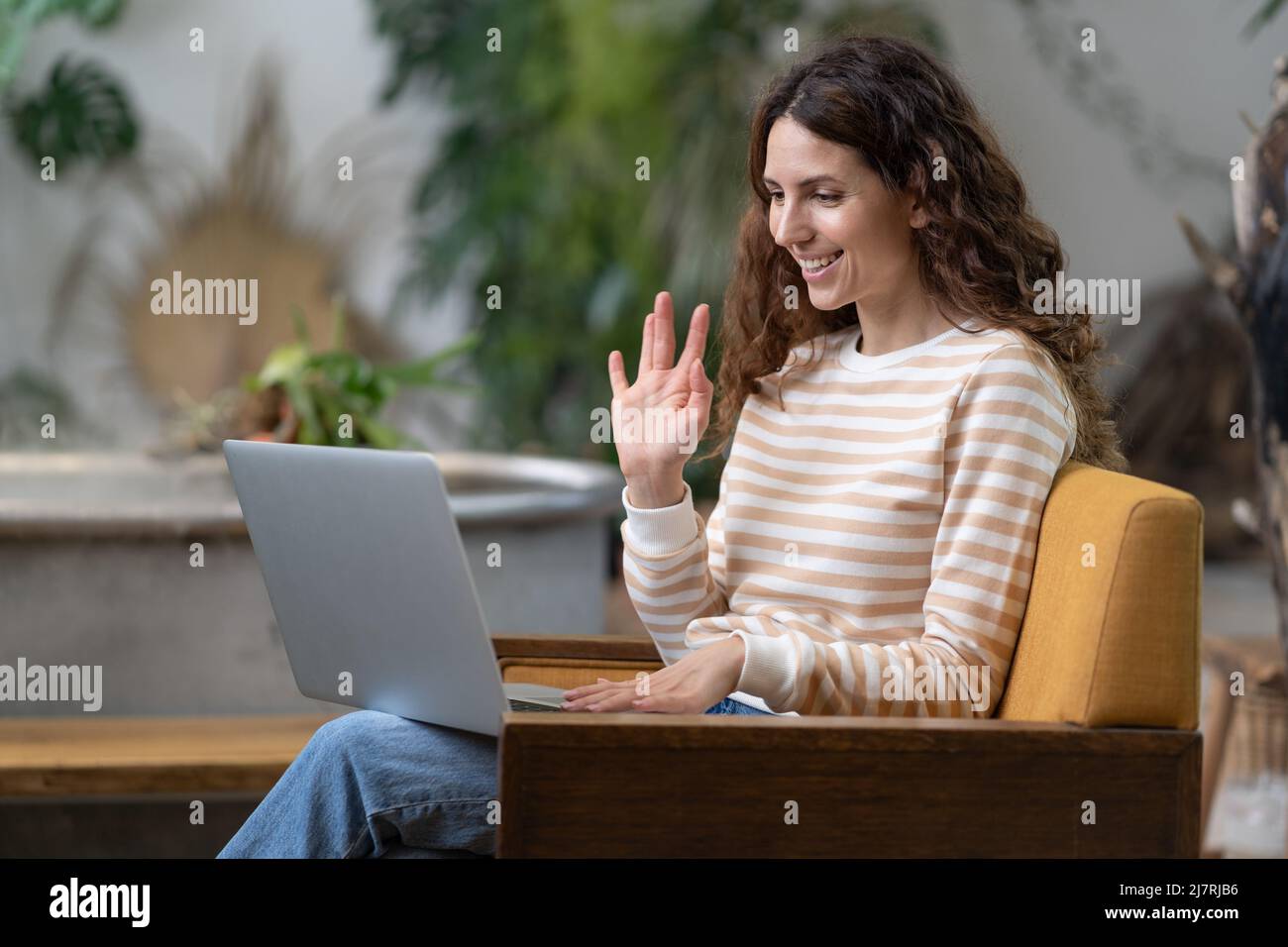 Donna italiana che sventola ciao, con video zoom chiamata su laptop seduta in poltrona in accogliente giardino di casa Foto Stock