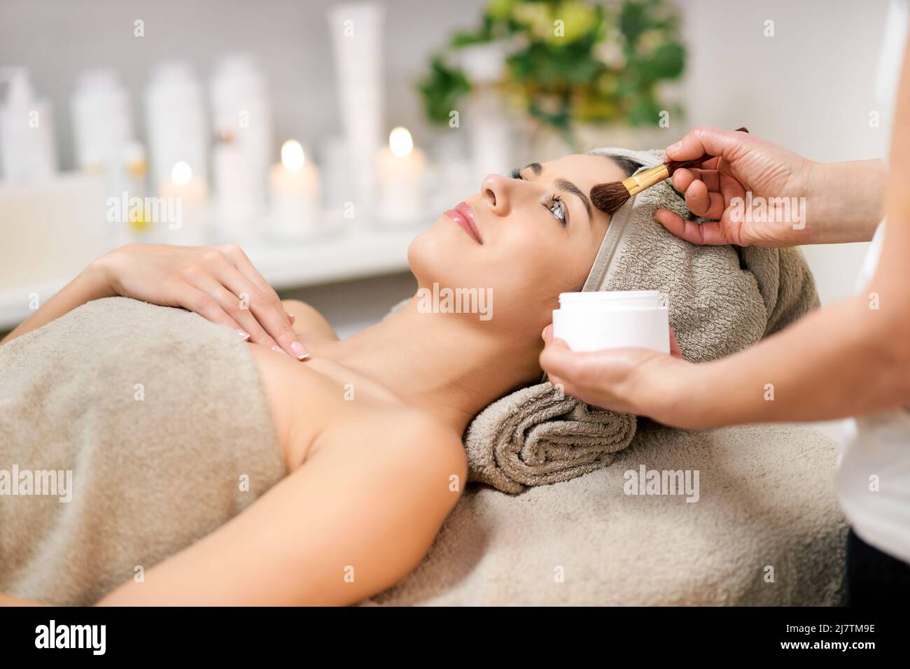 Crop anonymous cosmetician applicare il prodotto cosmetico sulla faccia del cliente femminile che si trova sotto l'asciugamano durante la procedura di bellezza nel salone termale. Concetto di make-up. Foto Stock