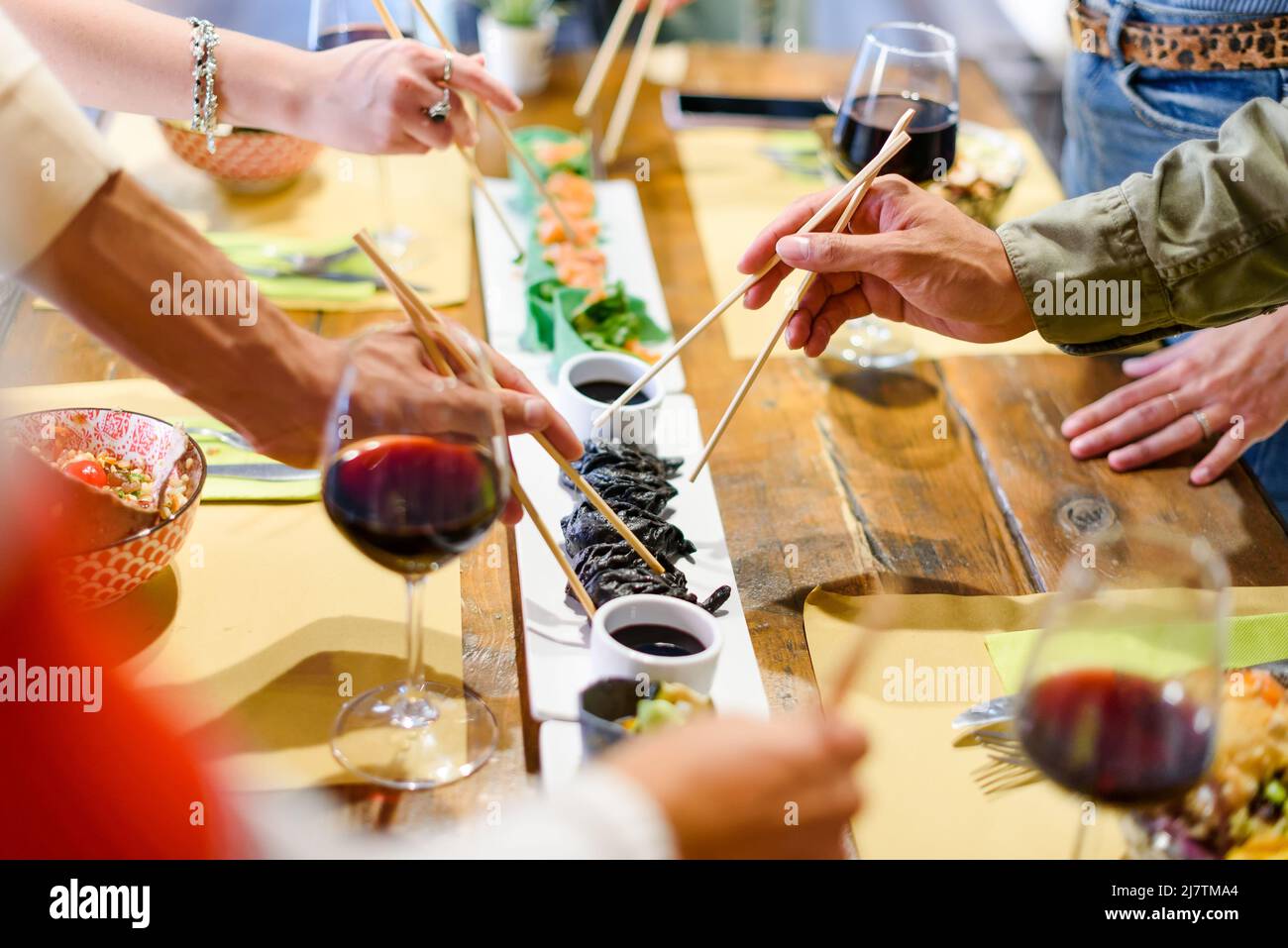 Alto angolo di raccolto anonimi amici che si riuniscono intorno al tavolo con piatti asiatici e bicchieri di vino rosso mentre si gusta la cena insieme Foto Stock