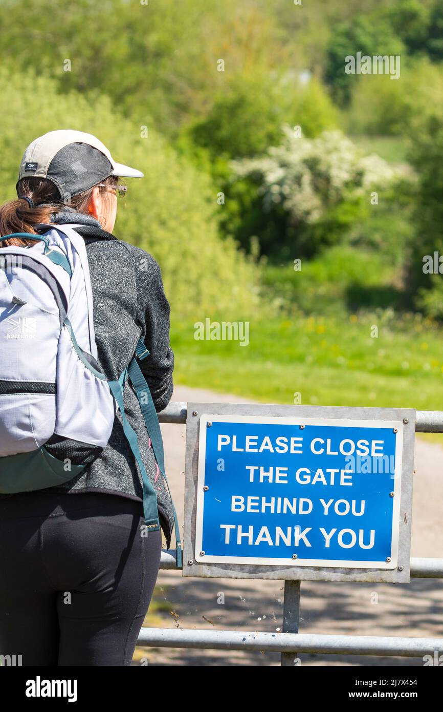 Vista posteriore di una donna escursionista sul sentiero del paese del Regno Unito, in piedi da un cartello gentile che dice "si prega di chiudere il cancello dietro di voi, grazie”. Foto Stock