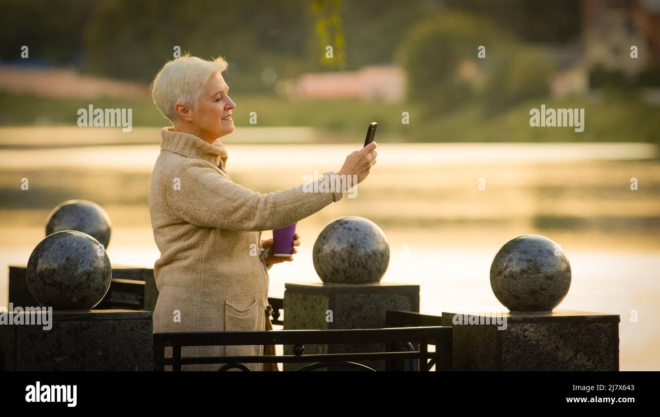 Signora di mezza età contented che fa la videochiamata guardando lo schermo dello smartphone che parla sul telefono delle cellule. Donna anziana in età di pensionamento scattando foto sopra Foto Stock