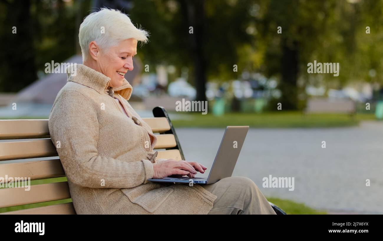 Donna anziana seduta su panchina nel parco della città utilizzando computer surf internet, sorridente anziana donna di età pensionabile che lavora a laptop leggere notizie Foto Stock