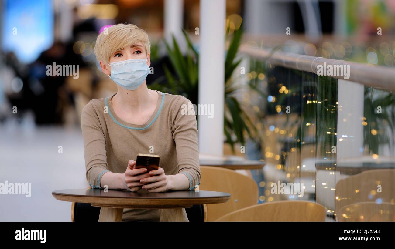 Donna caucasica adulta bionda in maschera medica matura femmina visitatore siede in caffetteria a tavola guarda il telefono controlla il tempo sullo smartphone guardando a lato Foto Stock