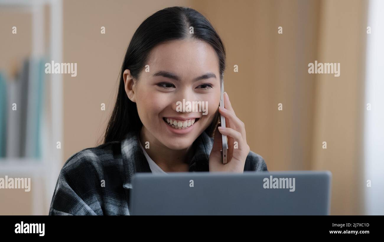 Felice sorridente ragazza asiatica coreana ragazza freelance femmina chattare con gli amici al telefono rispondere chiamata amichevole guardando il laptop schermo di lavoro remoto Foto Stock