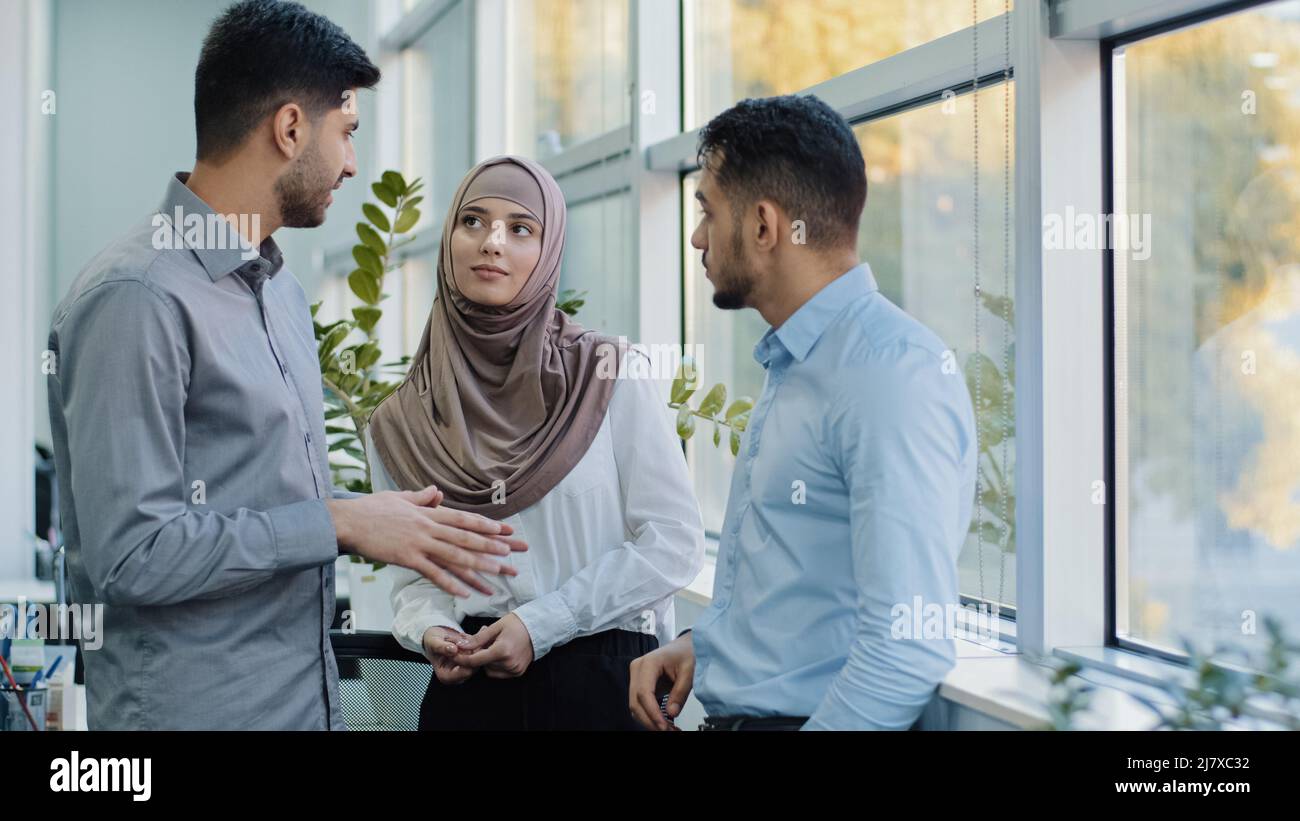 Colleghi multietnici la donna musulmana araba in hijab e due uomini millenni indiani discutono il compito aziendale. Lavoratori diversi uomini d'affari che lavorano Foto Stock