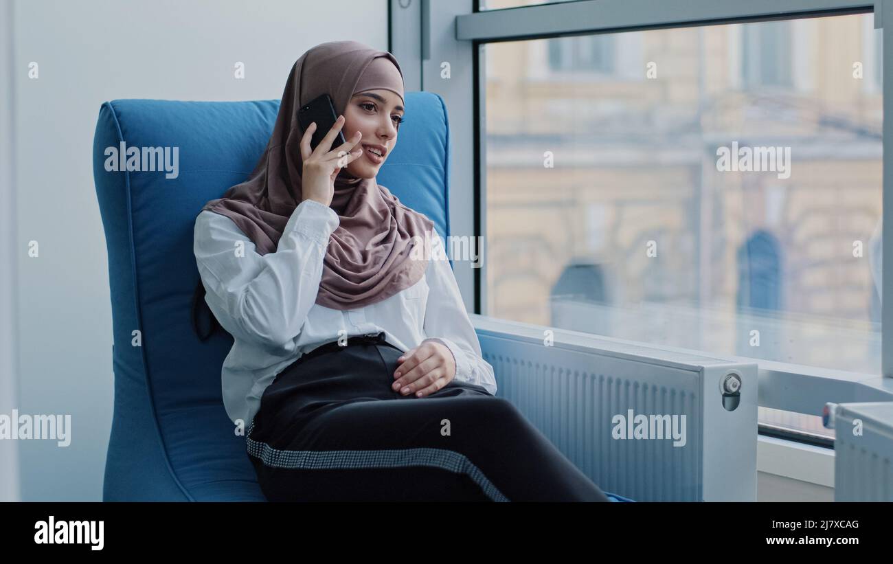 Sorridente ragazza araba rilassata manager millennial in hijab seduta in poltrona confortevole parlare al telefono, consulenza, fare business call al lavoro Foto Stock