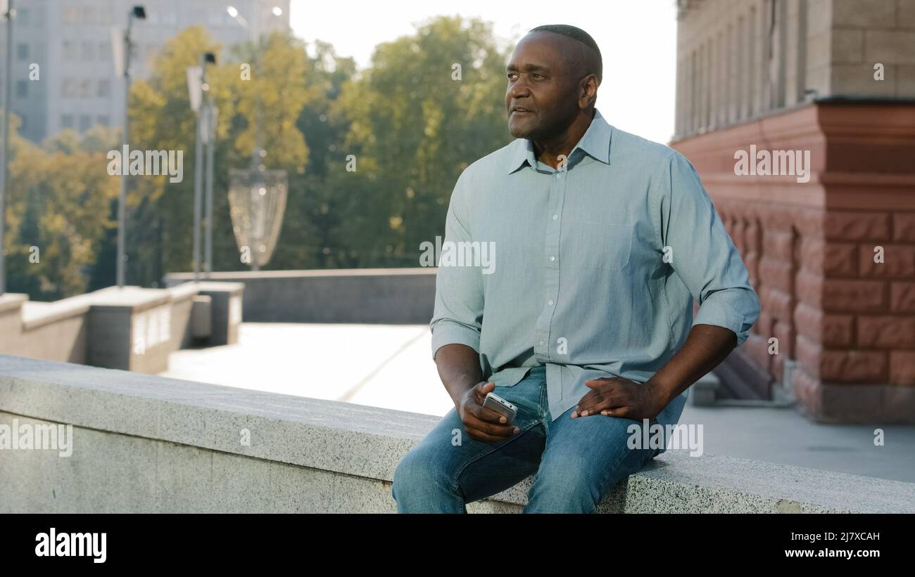 Stressato e frustrato l'uomo afro-americano di età pensionabile in abiti casual camicia e jeans seduti all'aperto tenendo telefono in attesa del cliente Foto Stock