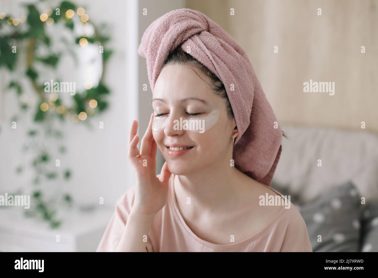 Giovane donna con gli occhi cerotti e asciugamano sulla testa a casa. Abitudini del mattino e la routine di bellezza domestica Foto Stock