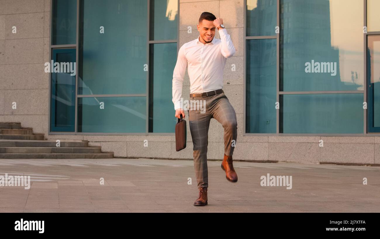 Vista a tutta lunghezza arabic spaniard uomo d'affari leader manager tenendo il laptop in valigetta in piedi all'aperto facendo vittoria gesto vince gioie Foto Stock