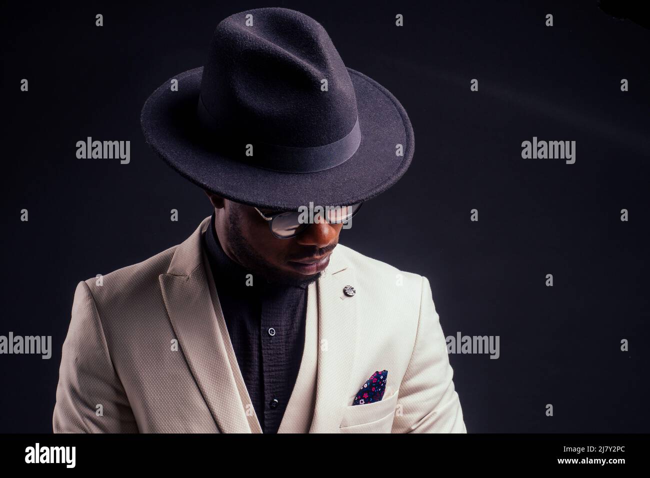 giovane bel ragazzo afro americano malinconico in una giacca bianca e occhiali su sfondo nero in studio.mistero e dolore Foto Stock