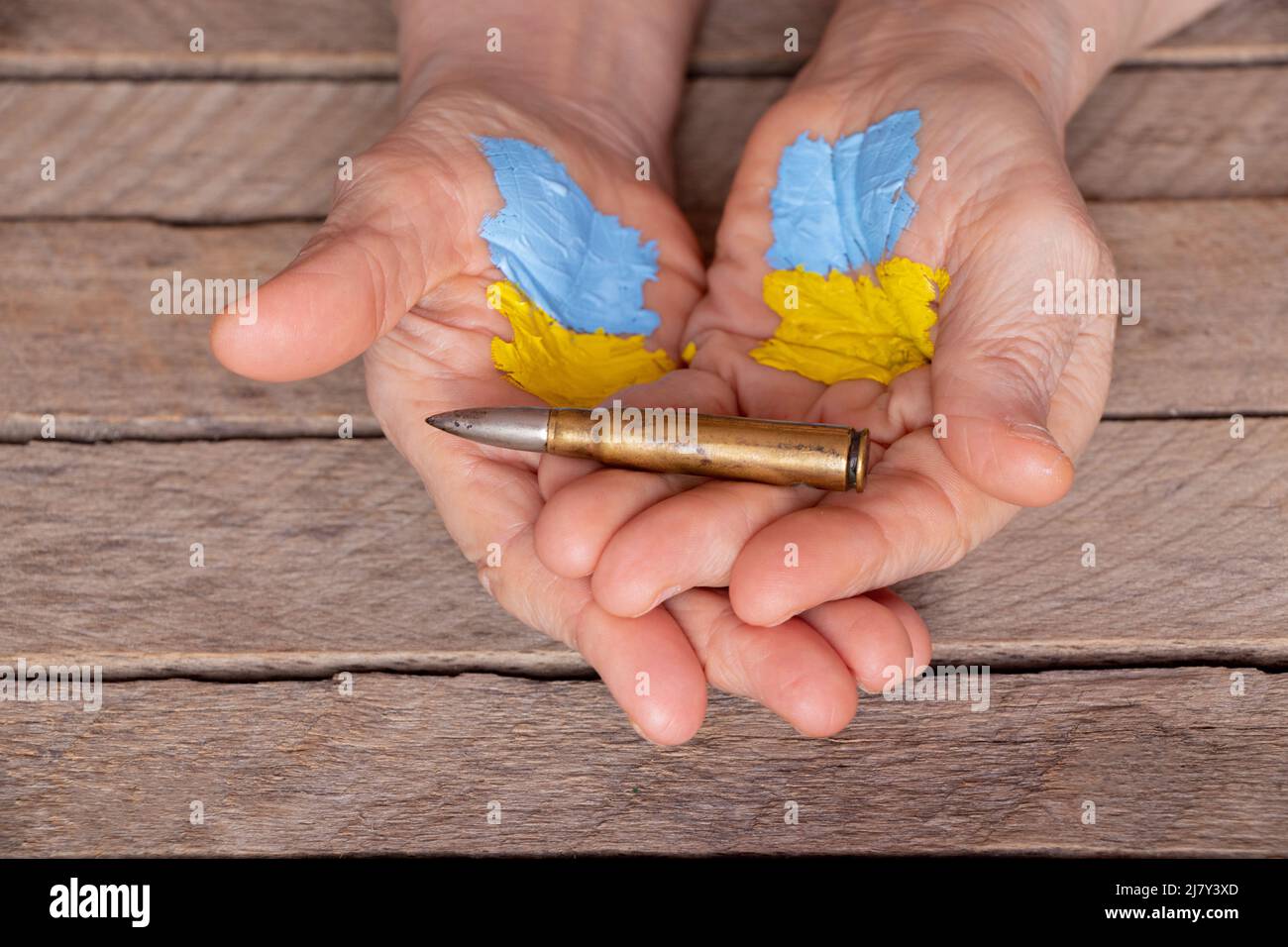 Le mani di una vecchia donna con una bandiera dipinta dell'Ucraina detengono una pallottola militare su uno sfondo di legno, guerra in Ucraina, donna militare Foto Stock