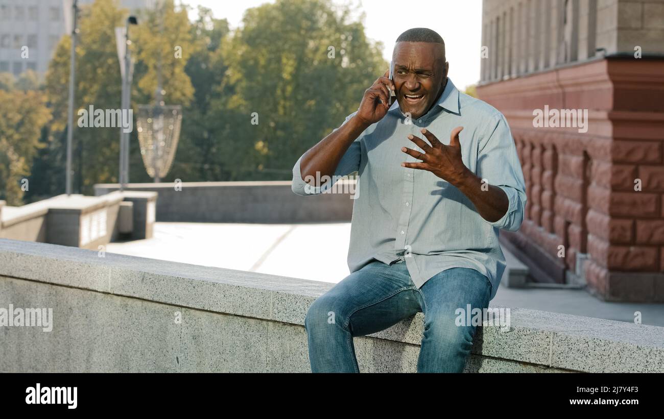 Preoccupato nervoso africano americano uomo in camicia e jeans che tiene il telefono cellulare. Cliente stressato frustrato dell'età di pensionamento che parla al servizio seduta Foto Stock
