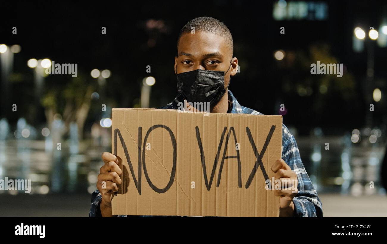Giovane afroamericano arrabbiato in maschera nera si trova in città la sera tenendo bandiera segno con testo No VAX Guy protesta del vaccino covid 19 Foto Stock