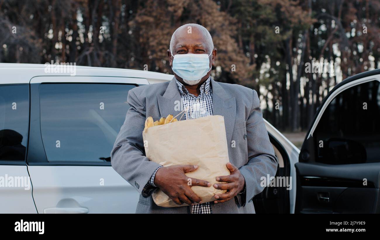 L'uomo africano Americano anziano in maschera medica protettiva nel parcheggio di strada ha comprato i generi alimentari in negozio di frutta anziano stativo maschio vicino ai supporti dell'automobile Foto Stock