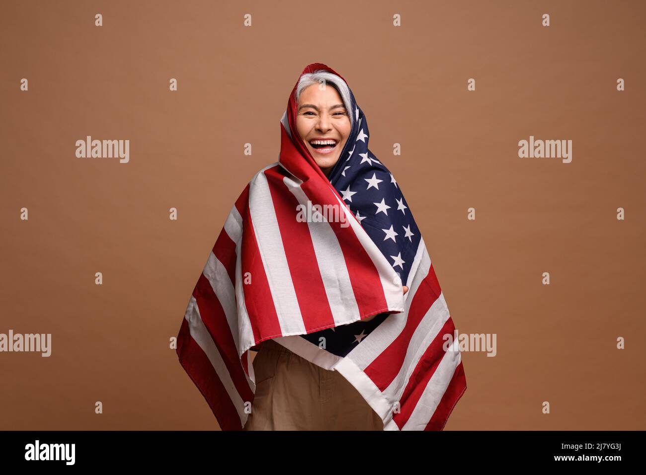 Ritratto di donna matura che avvolge la bandiera degli Stati Uniti isolato su sfondo marrone. La signora patriottica anziana, con grande gioia, festeggia il 4th luglio Foto Stock