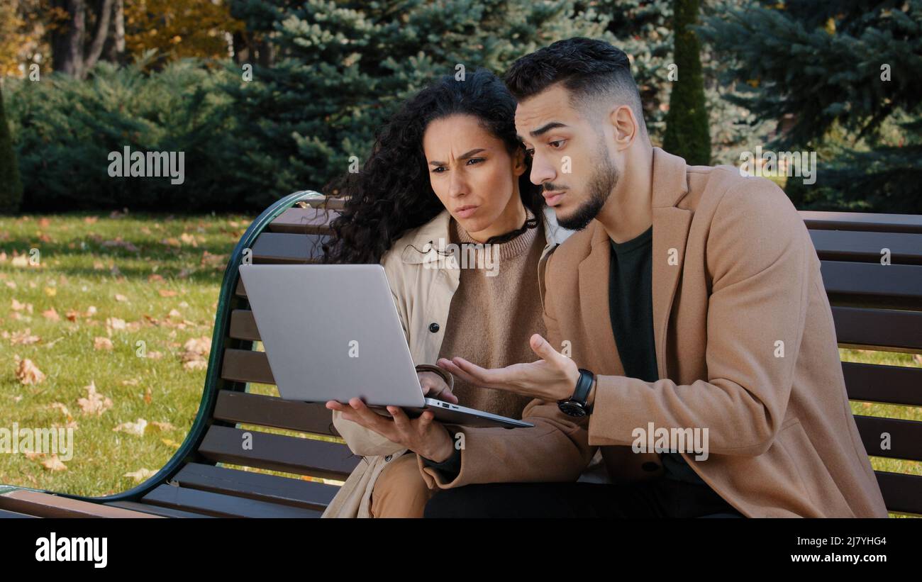 Preoccupato ispanico giovane coppia con laptop sedersi in panchina in autunno parco non riuscito shopping online rotto dispositivo serio frustrato partner commerciali Foto Stock