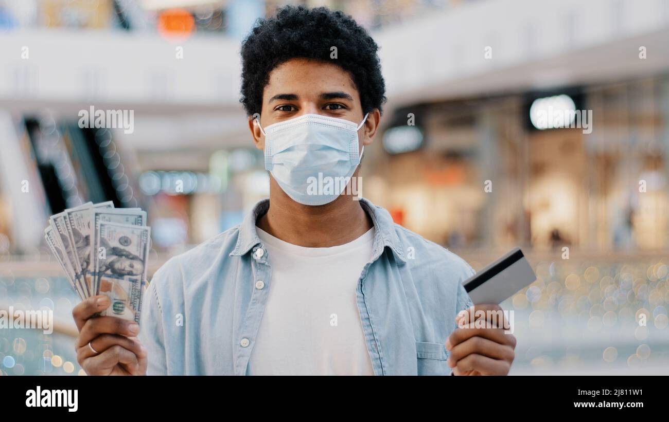 African american uomo uomo maschio vincitore indossa maschera medica viso tiene carta di credito e dollari in mani fa la scelta tra contanti e elettronica Foto Stock
