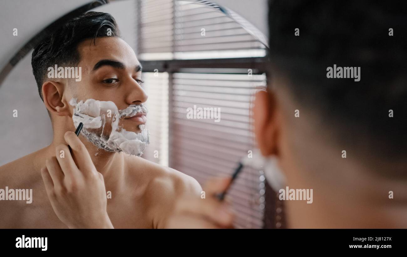 Ispanico arabo indiano bico bearded bel uomo rasatura usando il rasoio usa e getta in bagno guardando lo specchio. Giornaliero mattina di routine maschile igienico Foto Stock