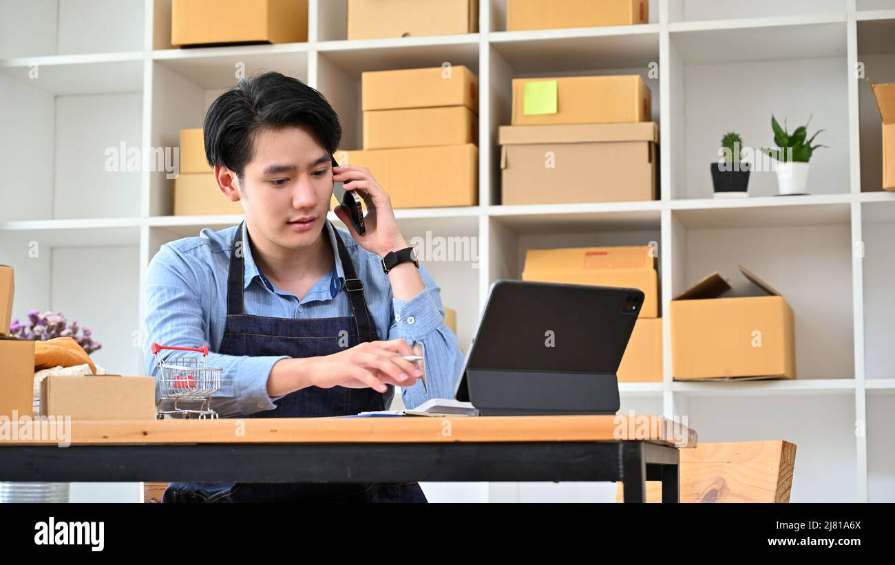 Il proprietario di affari del millennial asiatico intelligente del commercio di partenza del maschio che parla al telefono con il suo fornitore del materiale mentre lavora alla sua scrivania. Foto Stock