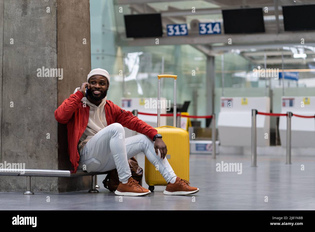 Sorridente giovane hipster afroamericano con valigia che chiama al telefono in attesa di volo in aeroporto. Foto Stock