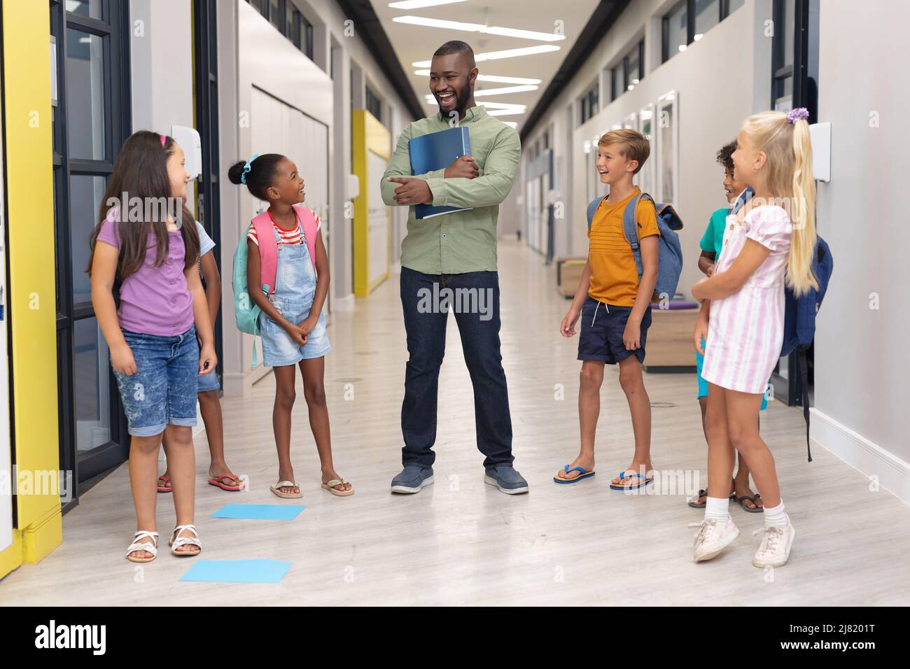 Giovane insegnante africano americano di sesso maschile che parla con studenti elementari multirazziali in piedi in corridoio Foto Stock