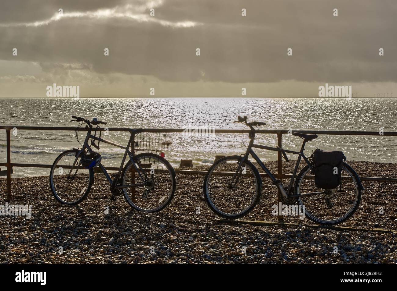 Due biciclette appoggiate su ringhiere su una spiaggia di ghiaia con cielo tempestoso e mare scintillante. Shoreham nel Sussex occidentale, Inghilterra Foto Stock