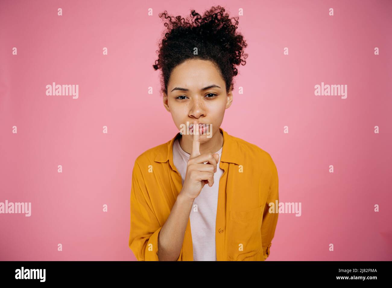 Gesto di silenzio, concetto segreto. Bella ragazza afroamericana in abbigliamento casual, mostrando segno shh con il dito vicino alle labbra, gesto di silenzio, in piedi su sfondo rosa isolato, guardando la macchina fotografica Foto Stock