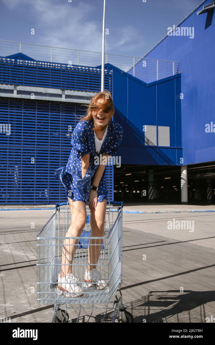 Una giovane donna in occhiali da sole e un vestito blu rotola intorno in un carrello della drogheria contro lo sfondo di un centro commerciale blu o di un supermercato. Foto Stock