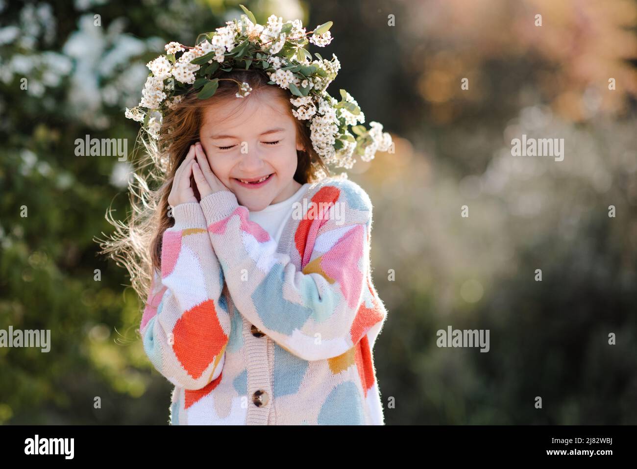 Felice sorridente ragazza 6-7 anni indossare maglia colorato maglione e floreale wreath acconciatura con fiori in parco sullo sfondo della natura. Primavera. SP Foto Stock