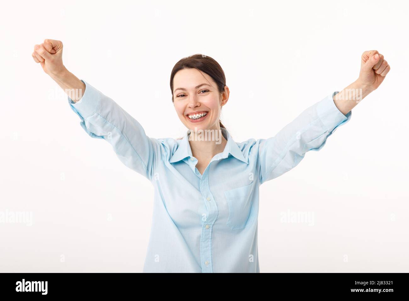 Stupito brunette giovane donna d'affari in camicia casual è gesturing vittoria con le sue mani sollevate, è scioccata, estremamente felice, sorridere fascio, sopra Foto Stock