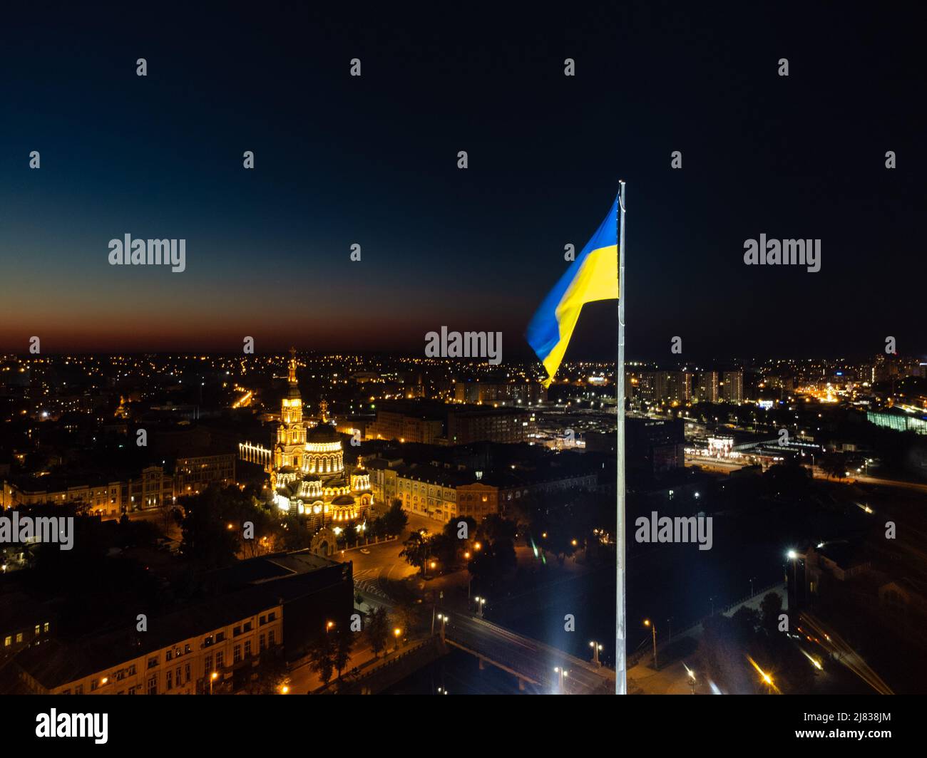 Sorvola la bandiera illuminata gialla e blu dell'Ucraina ondeggiando sul flagpole di notte. Vista aerea delle luci della città con la Cattedrale della Santa Annunciazione a Kharki Foto Stock