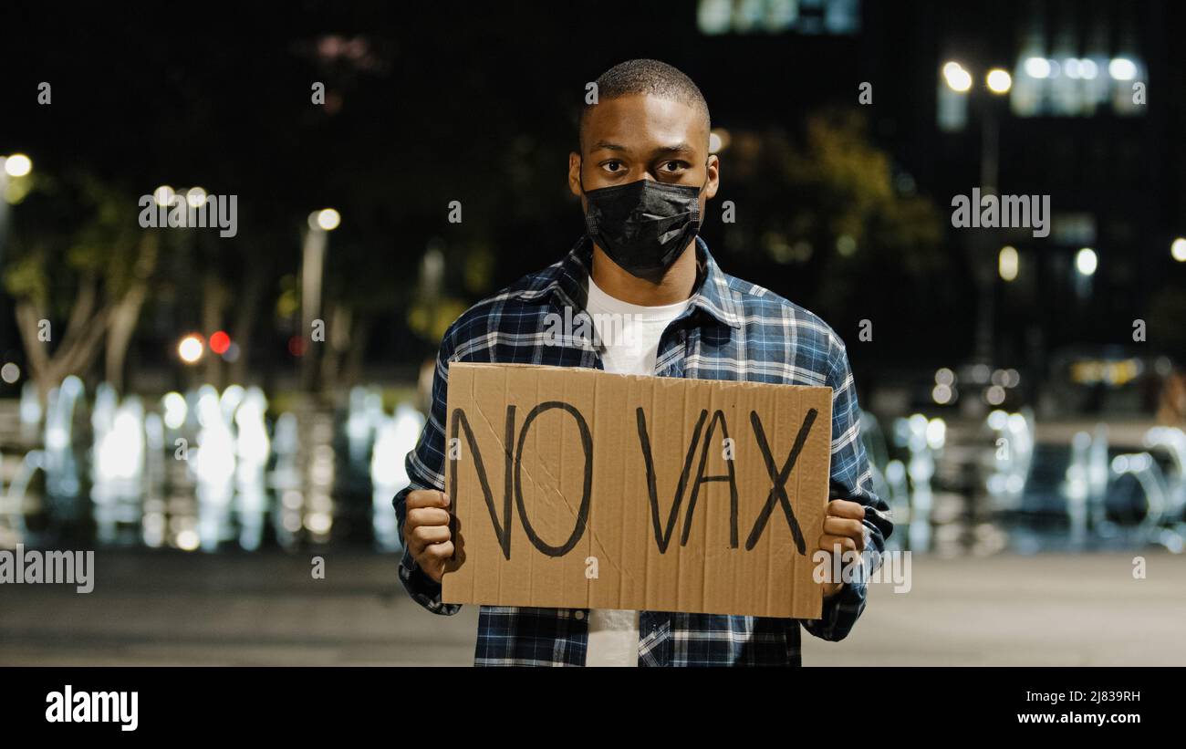 Ritratto afroamericano arrabbiato uomo in maschera di faccia si erge in città di notte sera tenendo segno banner con testo No VAX Guy protesta di covid 19 vaccino Foto Stock