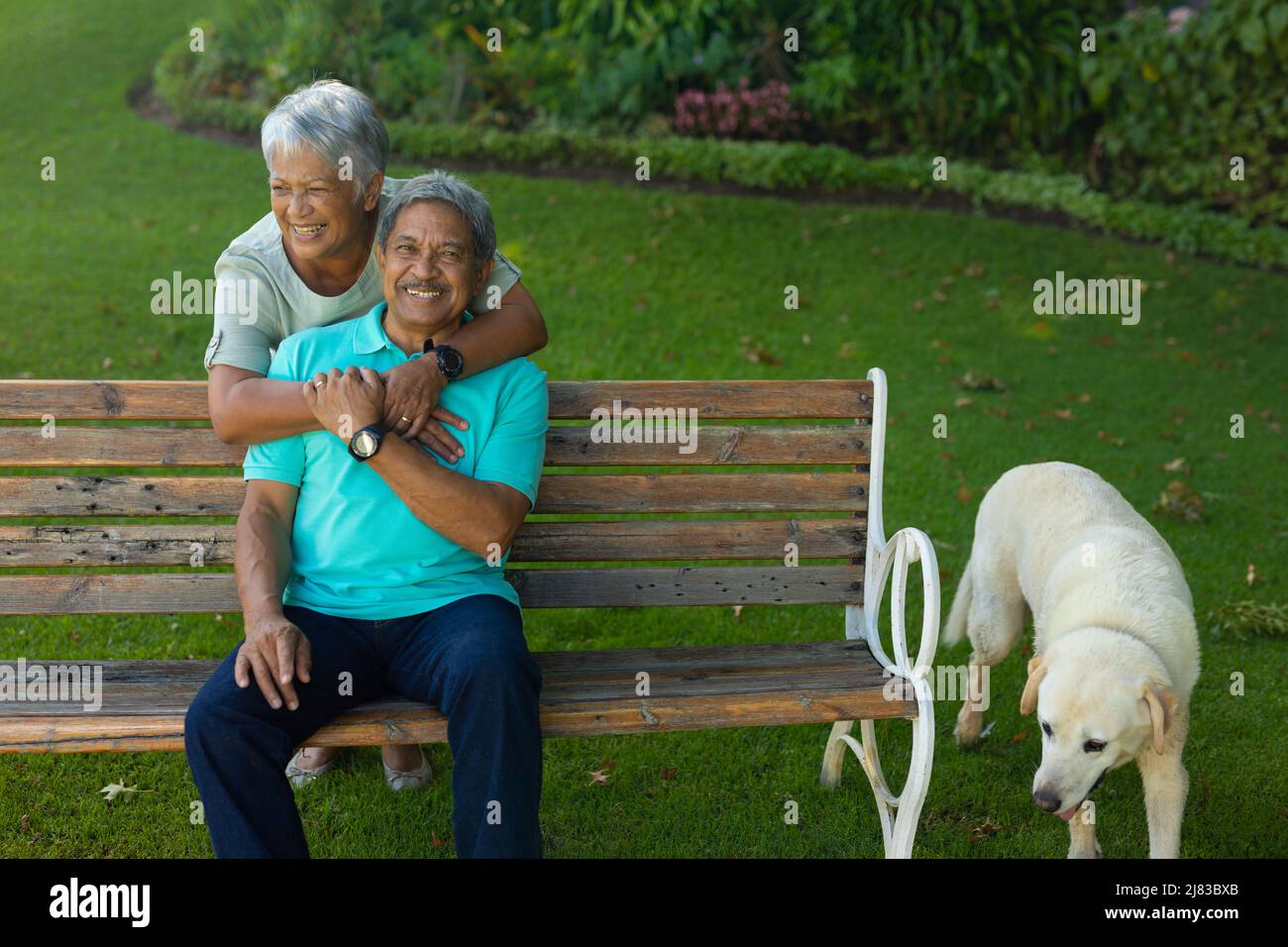 Cane in piedi da panca e ragazza allegra biraciale che abbraccia marito seduto su panca nel parco Foto Stock