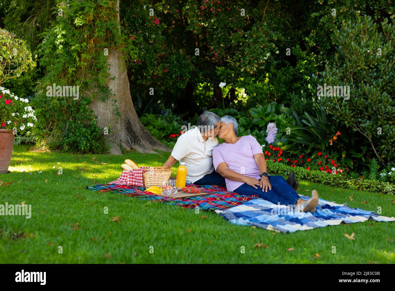Coppia anziana biraciale baciare mentre si siede con cibo e bevande su coperta contro le piante nel parco Foto Stock