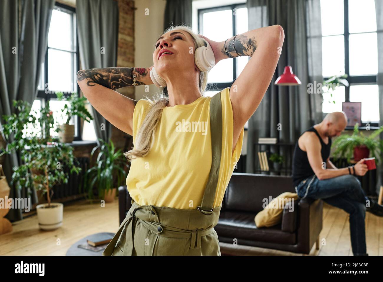 Donna caucasica matura ed elegante con tatuaggi sulle braccia che si godono la musica in cuffia, suo marito bere caffè su sfondo Foto Stock