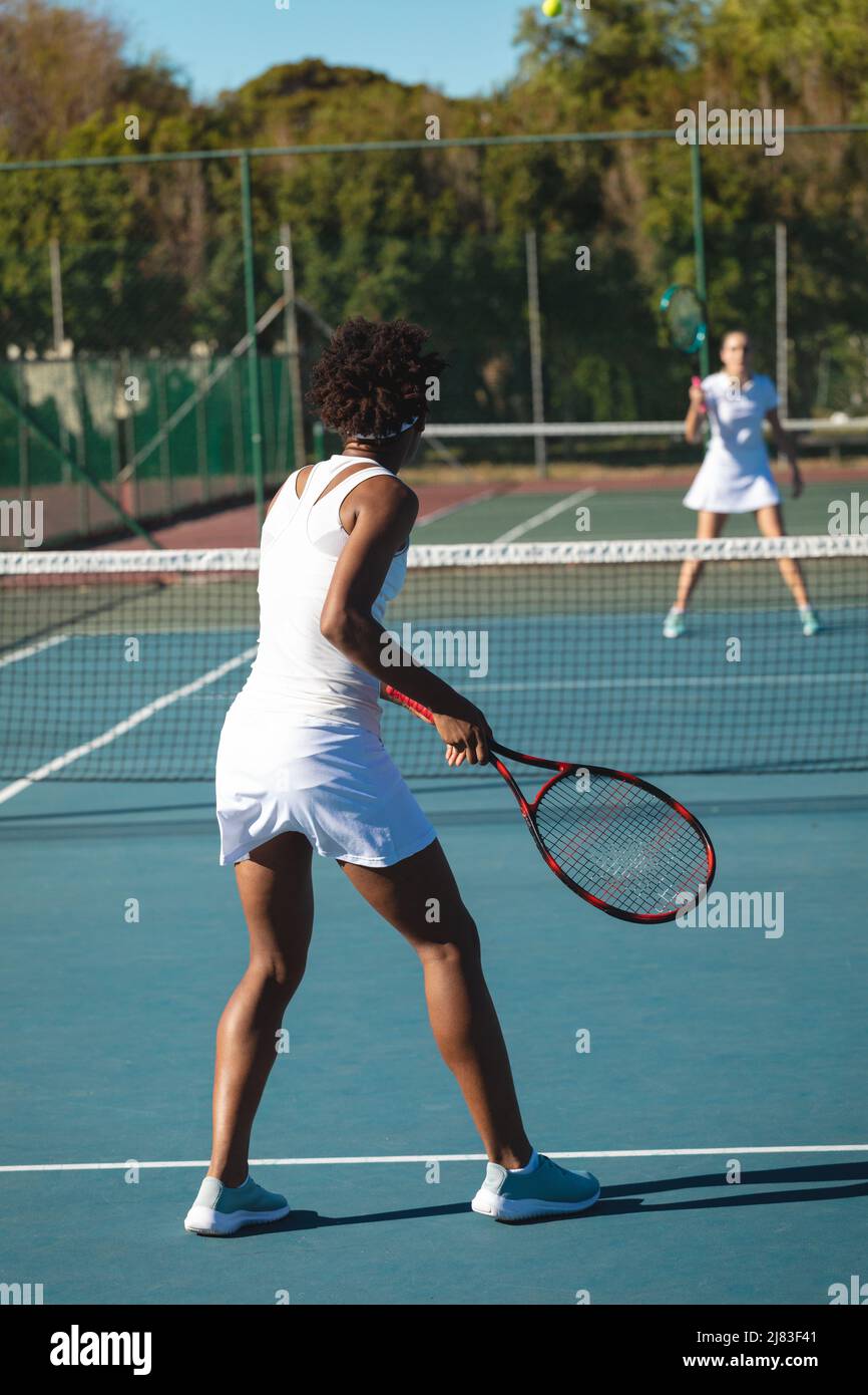 Una donna afro-americana che gioca a tennis con un concorrente caucasico in campo il giorno di sole Foto Stock