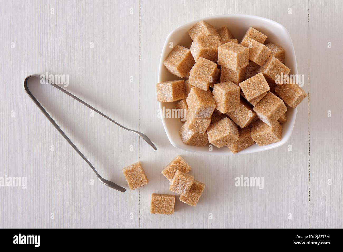Zucchero di canna a forma di cubo in ciotola di legno su un tavolo di legno bianco con pinze di zucchero d'argento. Piatto, vista dall'alto, niente persone. Foto Stock