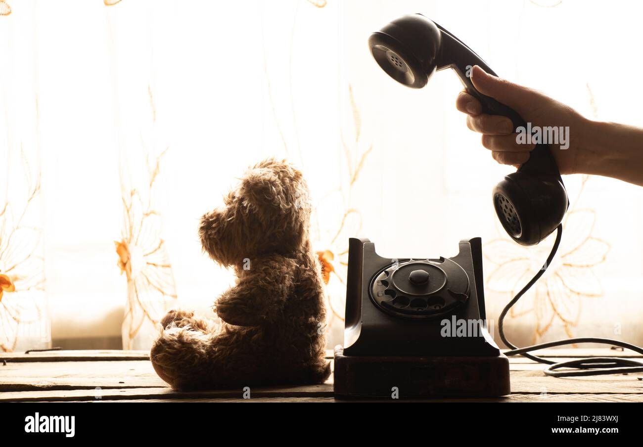 La mano di una donna compone un numero di telefono su un vecchio telefono e un orsacchiotto si siede accanto ad esso a casa sul tavolo, un telefono retrò è nero, una chiamata Foto Stock