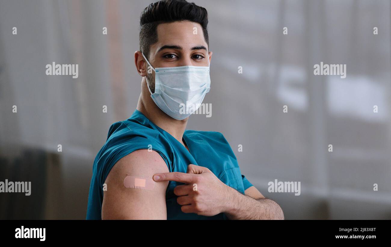 Uomo arabo medico infermiere indossare maschera chirurgica viso dimostrare segno di iniezione che punta alla spalla adesiva bendaggio dopo globale Foto Stock