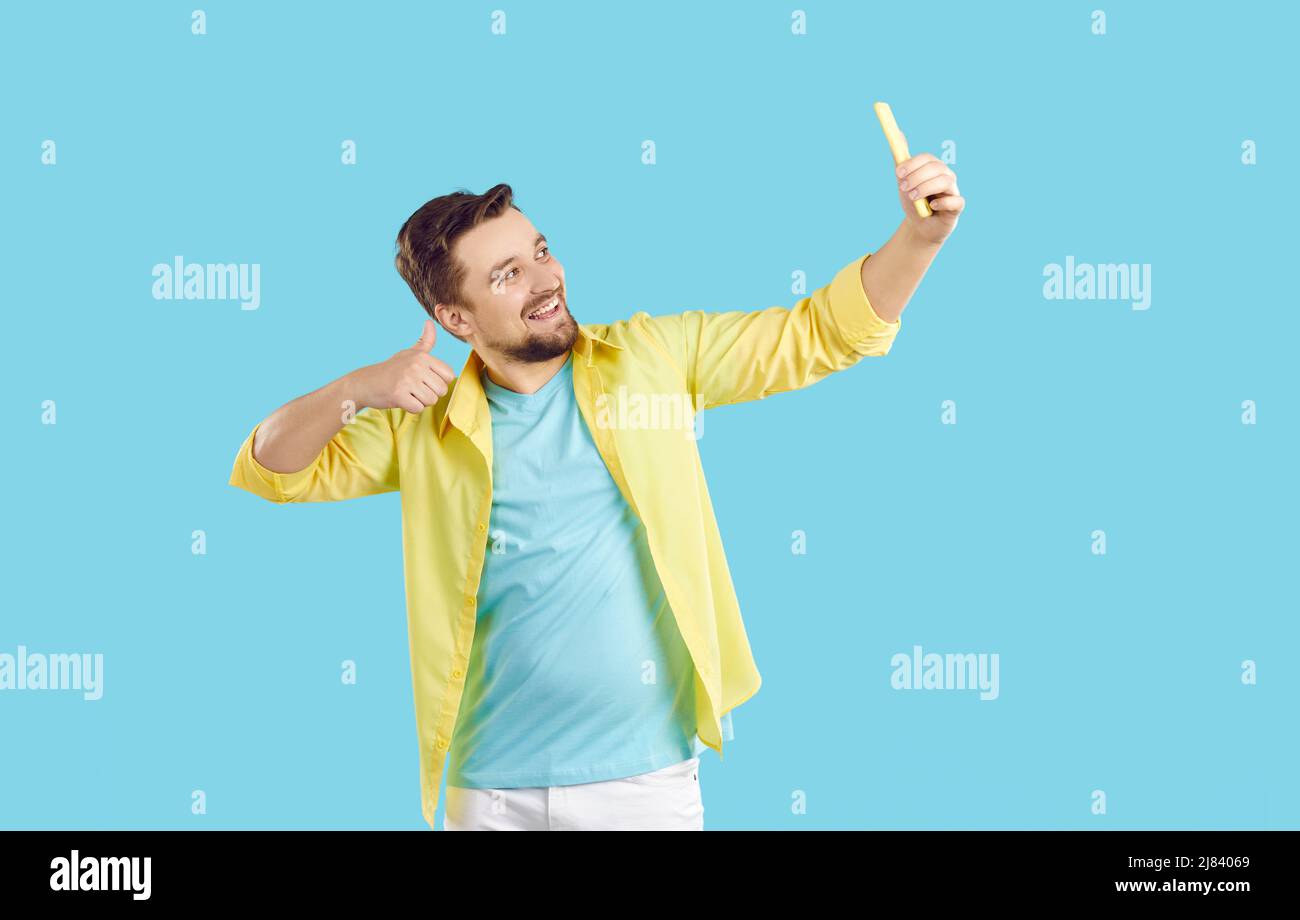 Felice uomo giovane sorridente, mostrando i pollici e prendendo selfie sul suo cellulare Foto Stock